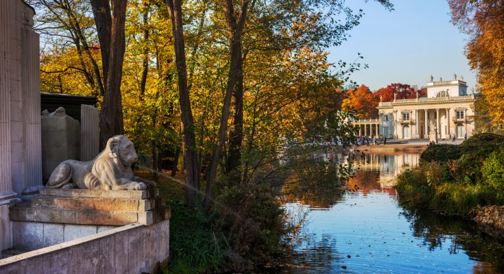 Herbstlicher Łazienki-Park mit Palast auf der Insel, Löwenskulptur und ruhigem Wasser in Warschau.