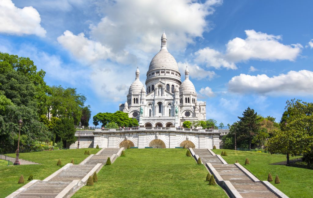 Weiße Kuppelbasilika Sacré-Cœur auf dem Montmartre-Hügel mit breiten Treppen und grüner Wiese unter blauem Himmel.