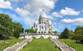 Weiße Kuppelbasilika Sacré-Cœur auf dem Montmartre-Hügel mit breiten Treppen und grüner Wiese unter blauem Himmel.