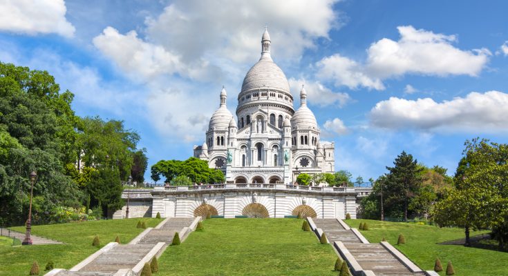 Weiße Kuppelbasilika Sacré-Cœur auf dem Montmartre-Hügel mit breiten Treppen und grüner Wiese unter blauem Himmel.