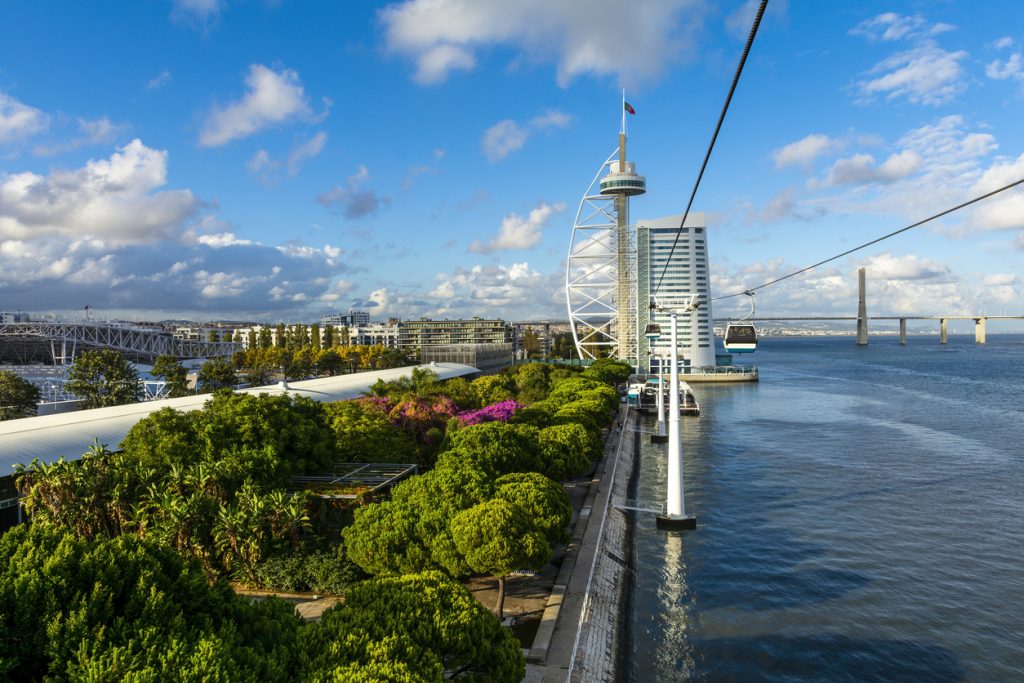 Moderne Flusslandschaft in Lissabon mit schwebender Seilbahn über dem Tejo, dem weißen Vasco-da-Gama-Turm/Hotel und der Vasco-da-Gama-Brücke im Hintergrund.
