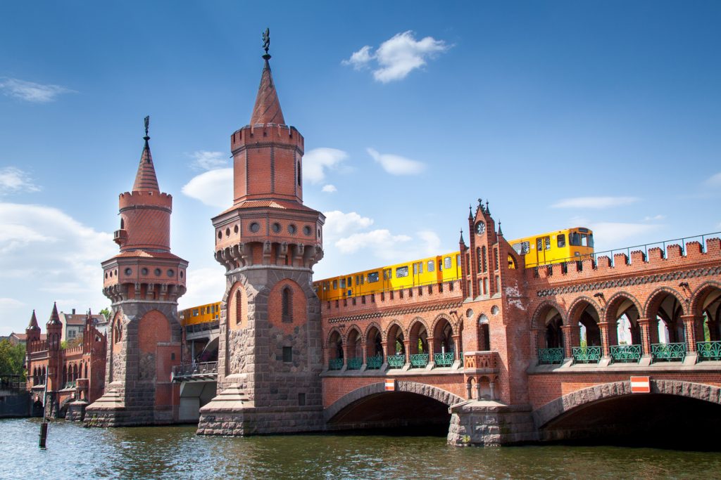 Rote Backstein-Oberbaumbrücke mit zwei neogotischen Türmen über der Spree; darüber fährt eine gelbe U-Bahn, blauer Himmel, Wasser im Vordergrund.