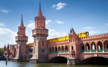 Rote Backstein-Oberbaumbrücke mit zwei neogotischen Türmen über der Spree; darüber fährt eine gelbe U-Bahn, blauer Himmel, Wasser im Vordergrund.