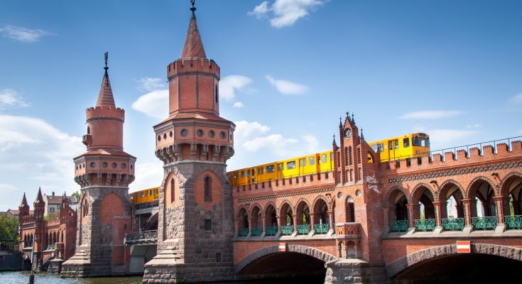 Rote Backstein-Oberbaumbrücke mit zwei neogotischen Türmen über der Spree; darüber fährt eine gelbe U-Bahn, blauer Himmel, Wasser im Vordergrund.