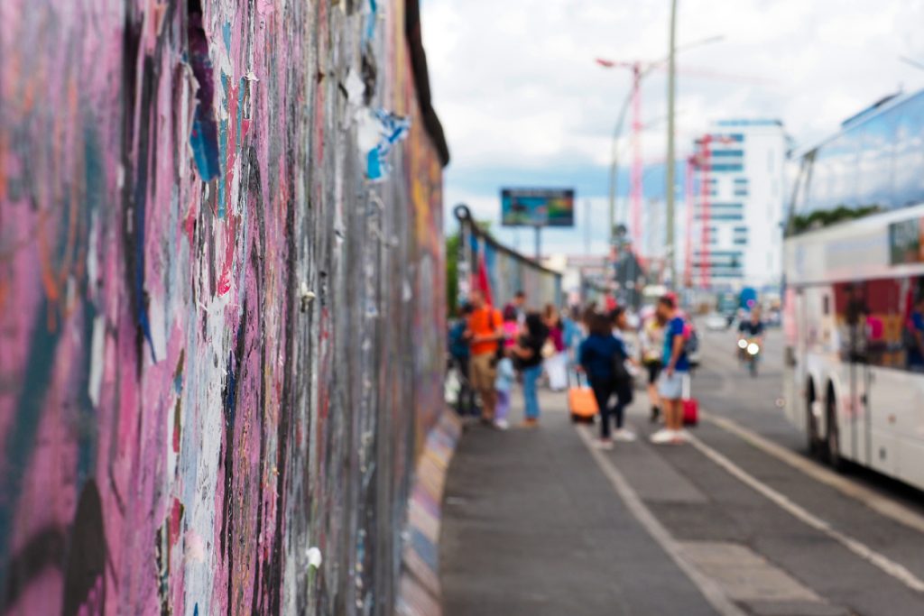 Nahaufnahme der bunt bemalten Berliner Mauer an der East Side Gallery; im unscharfen Hintergrund spazieren Besucher die Mühlenstraße entlang, rechts ein Bus.