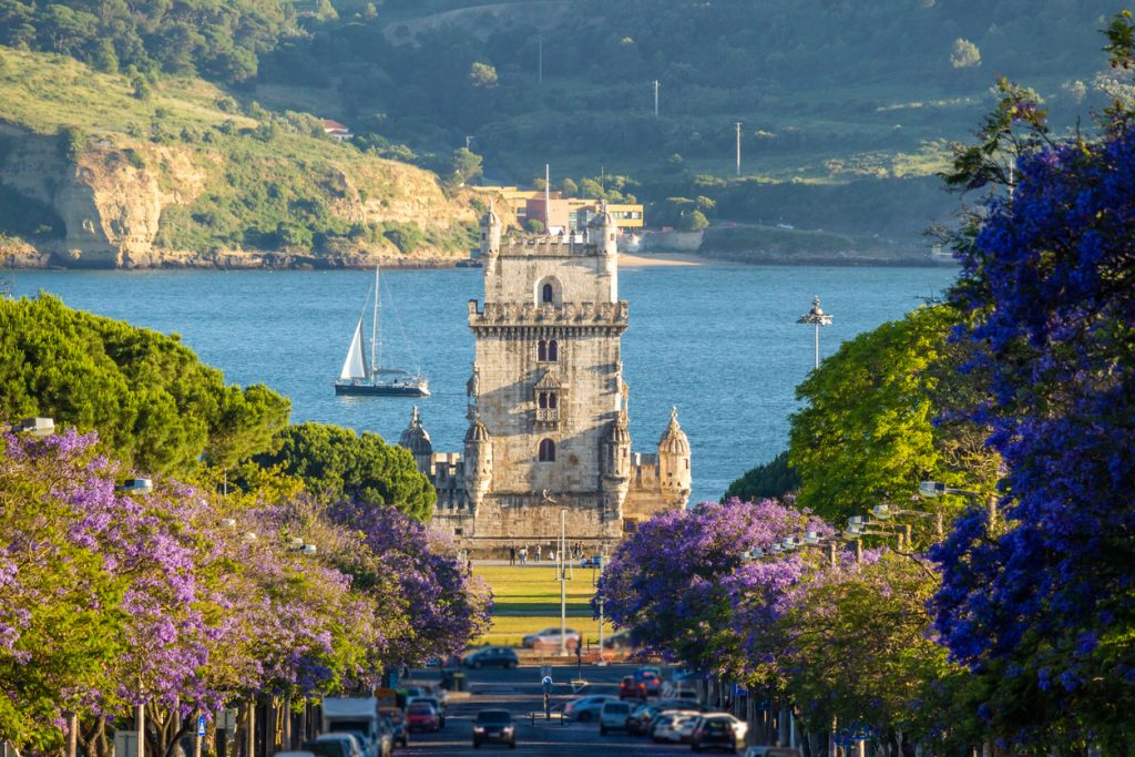 Jacarandagesäumte Allee mit Blick auf den Torre de Belém vor dem Tejo, ein Segelboot kreuzt im Hintergrund.
