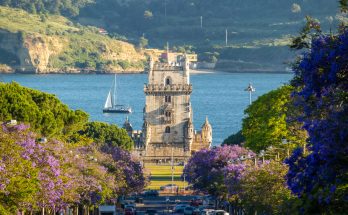 Jacarandagesäumte Allee mit Blick auf den Torre de Belém vor dem Tejo, ein Segelboot kreuzt im Hintergrund.
