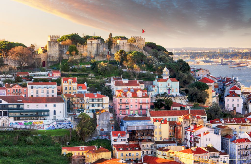 Hügel mit Burgmauern des Castelo de São Jorge über farbigen Alfama-Häusern, dahinter der Tejo unter Abendhimmel.