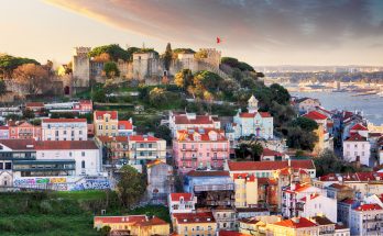Hügel mit Burgmauern des Castelo de São Jorge über farbigen Alfama-Häusern, dahinter der Tejo unter Abendhimmel.
