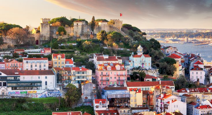Hügel mit Burgmauern des Castelo de São Jorge über farbigen Alfama-Häusern, dahinter der Tejo unter Abendhimmel.
