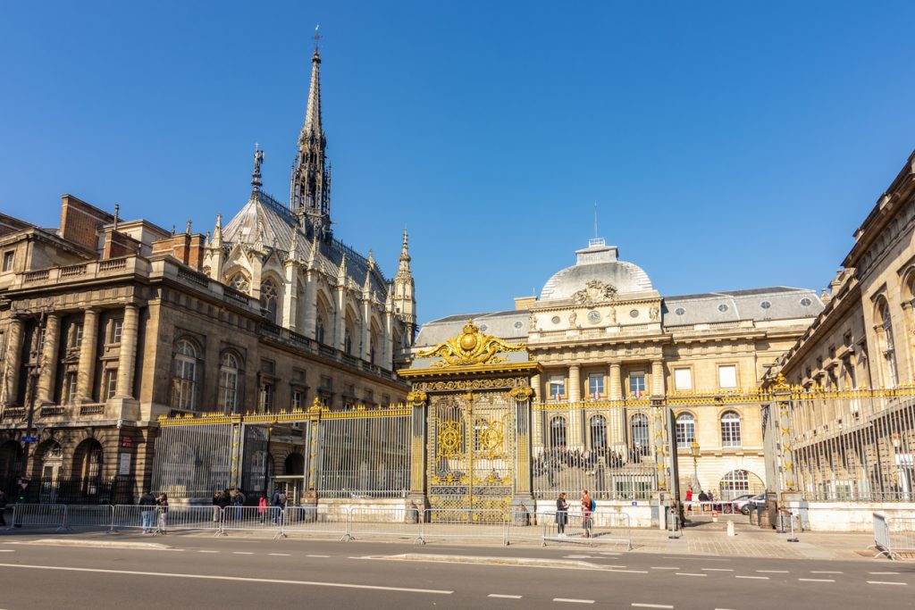 Gotische Sainte-Chapelle mit hohem Spitzhelm neben dem Palais de Justice, davor ein prunkvolles vergoldetes Tor unter blauem Himmel.