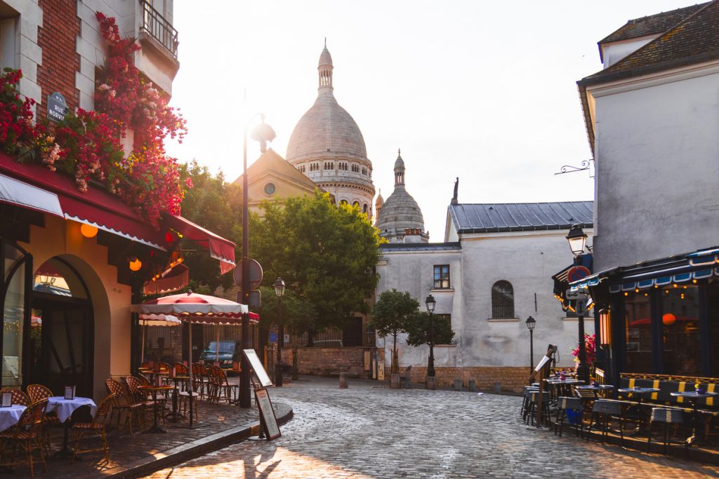 Pflasterstraße mit Straßencafés und roten Markisen in Montmartre, dahinter die weiße Kuppel der Basilika Sacré-Cœur im Gegenlicht.