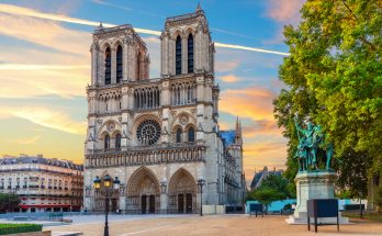 Fassade der Kathedrale Notre-Dame mit zwei Türmen und großer Rosette, rechts die Reiterstatue Karls des Großen unter farbigem Abendhimmel.
