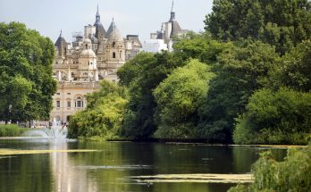 Parksee mit Springbrunnen und üppigen Bäumen, historische Gebäude im Hintergrund.