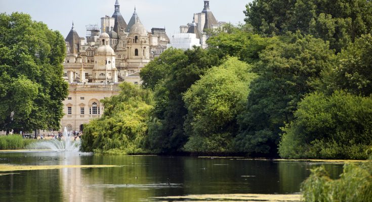 Parksee mit Springbrunnen und üppigen Bäumen, historische Gebäude im Hintergrund.