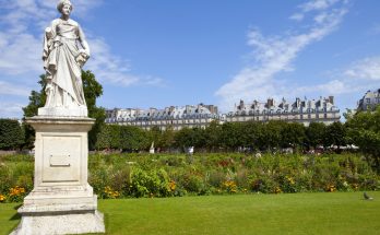 Marmorskulptur im Tuileriengarten vor blühenden Beeten und Haussmann-Dächern unter blauem Himmel.