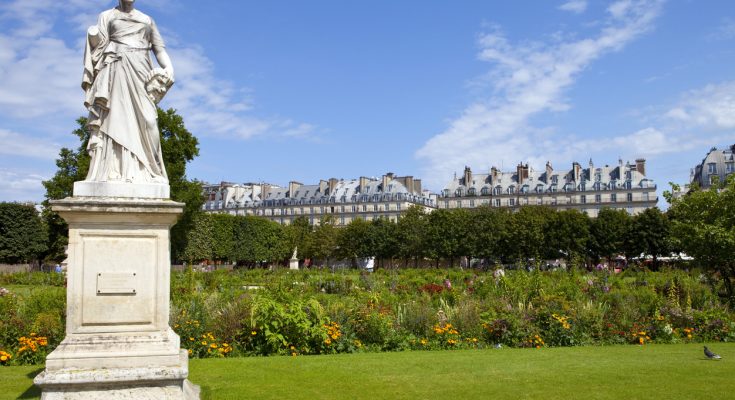 Marmorskulptur im Tuileriengarten vor blühenden Beeten und Haussmann-Dächern unter blauem Himmel.