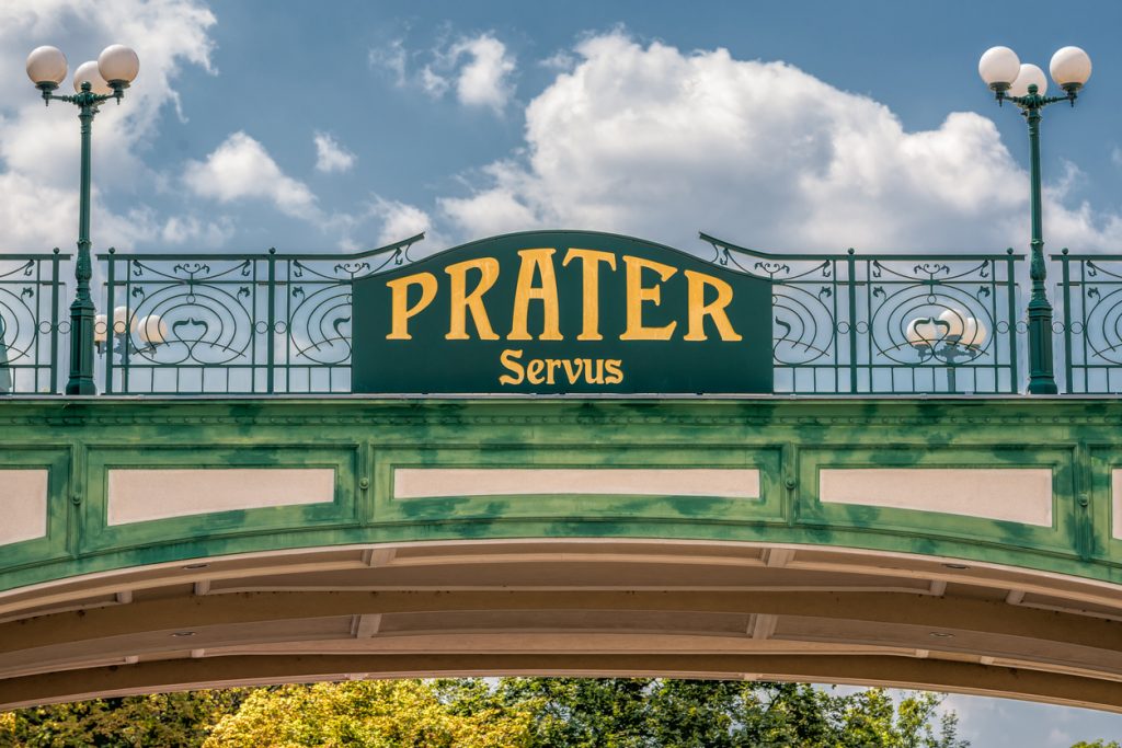 Grünes Schild „Prater – Servus“ mit Jugendstilgeländer und Lampen.