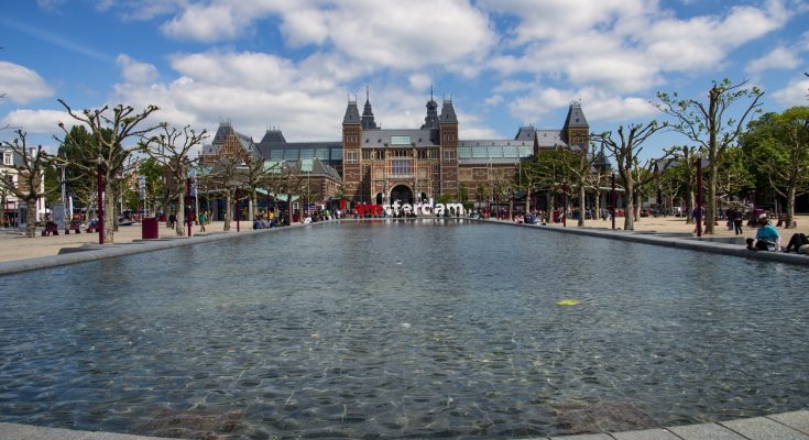 Frontansicht des Rijksmuseums mit langem Wasserbecken und Besuchern auf dem Museumplein.