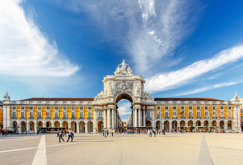 Weitläufiger Platz am Tejo mit gelben Arkadenbauten und dem weißen Triumphbogen Arco da Rua Augusta; Menschen flanieren über das Pflaster.