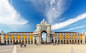 Weitläufiger Platz am Tejo mit gelben Arkadenbauten und dem weißen Triumphbogen Arco da Rua Augusta; Menschen flanieren über das Pflaster.