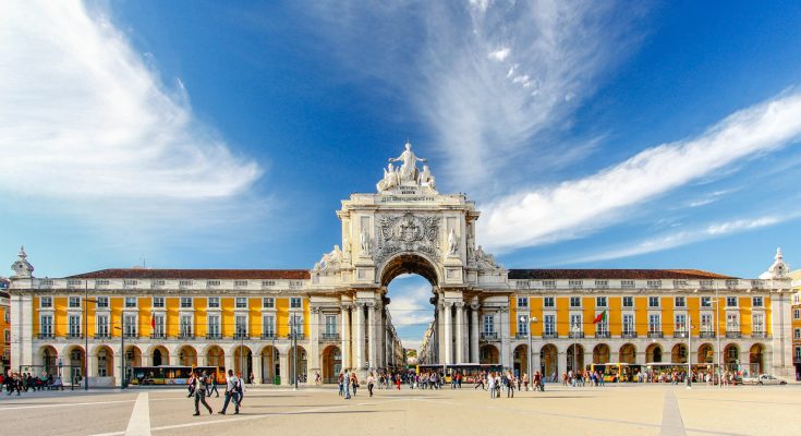 Weitläufiger Platz am Tejo mit gelben Arkadenbauten und dem weißen Triumphbogen Arco da Rua Augusta; Menschen flanieren über das Pflaster.