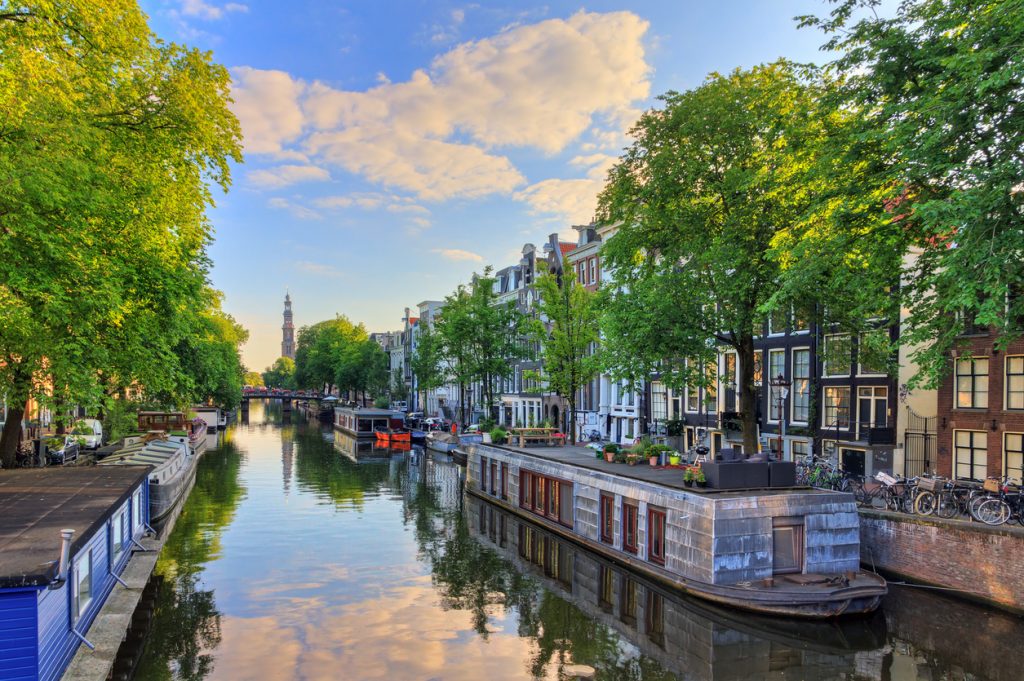 Blick von einer Brücke auf eine ruhige Gracht mit Hausbooten, giebeligen Grachtenhäusern und dem Turm der Westerkerk in der Ferne.
