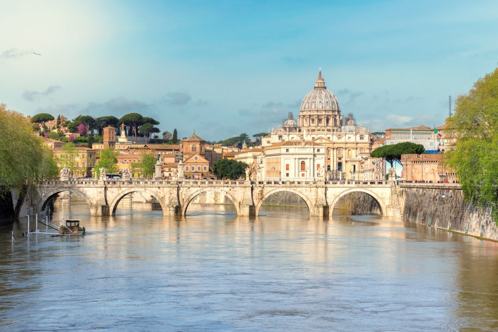 Blick über den Tiber auf den Petersdom mit historischer Steinbrücke im Vordergrund.