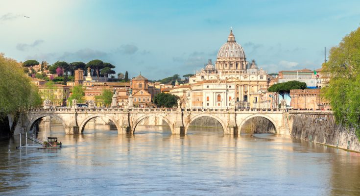 Blick über den Tiber auf den Petersdom mit historischer Steinbrücke im Vordergrund.